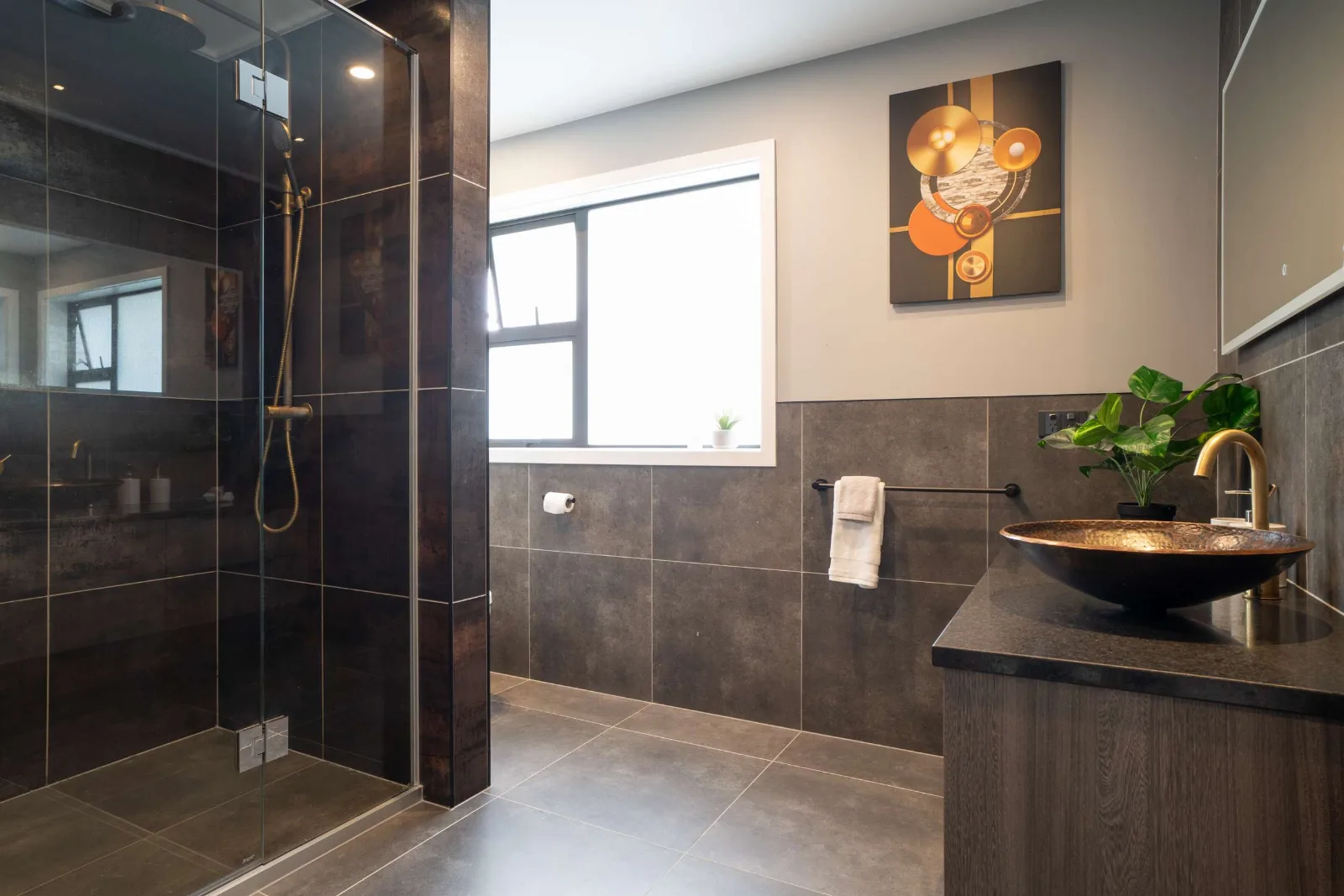 The tiled shower behind frameless glass — brass rail and rose, dark tile floor to ceiling, and the copper basin on its dark top to the right.