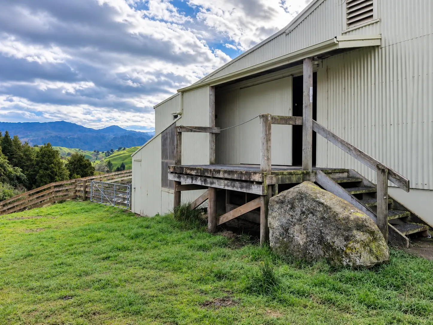 Taumarunui Woolshed — the end of the shed from the paddock, its timber loading platform standing off the corrugated wall with the hill country behind