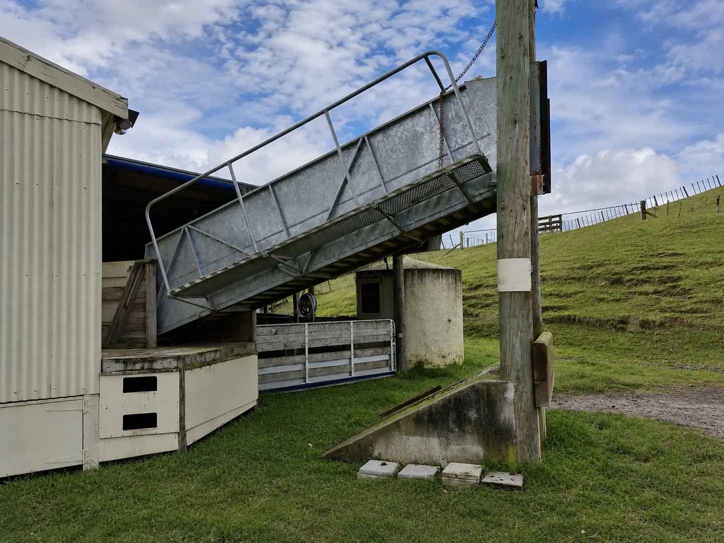The loading race from the yard — the galvanised race and its mesh walkway slung on a chain from the pole head, running down from the shed's loading deck to where a truck stands.